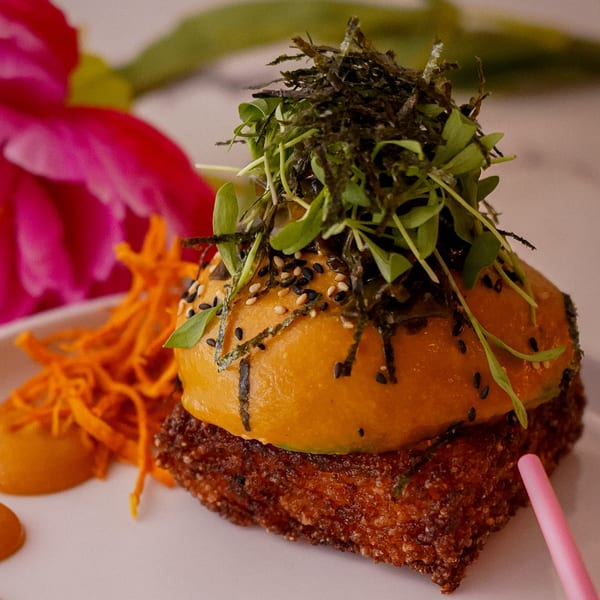 Vegan burger with orange bun, microgreens, seaweed, sesame seeds, and carrot strings on a white plate.