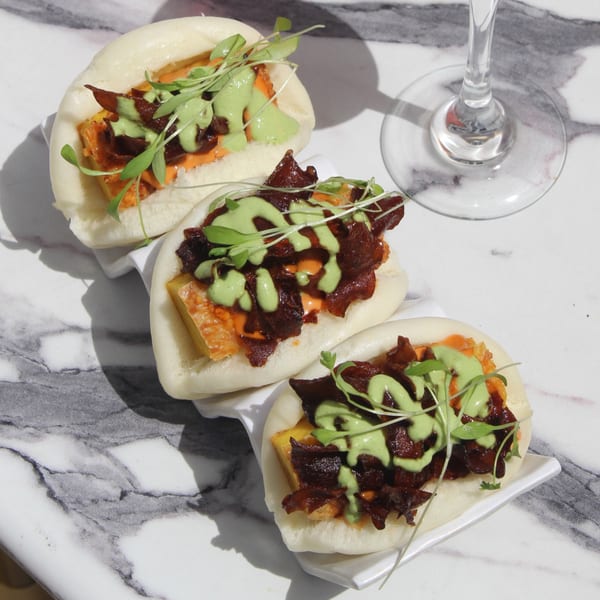 Three vegan bao buns with crispy tofu, dark sauce, green drizzle, and microgreens on a marble table.